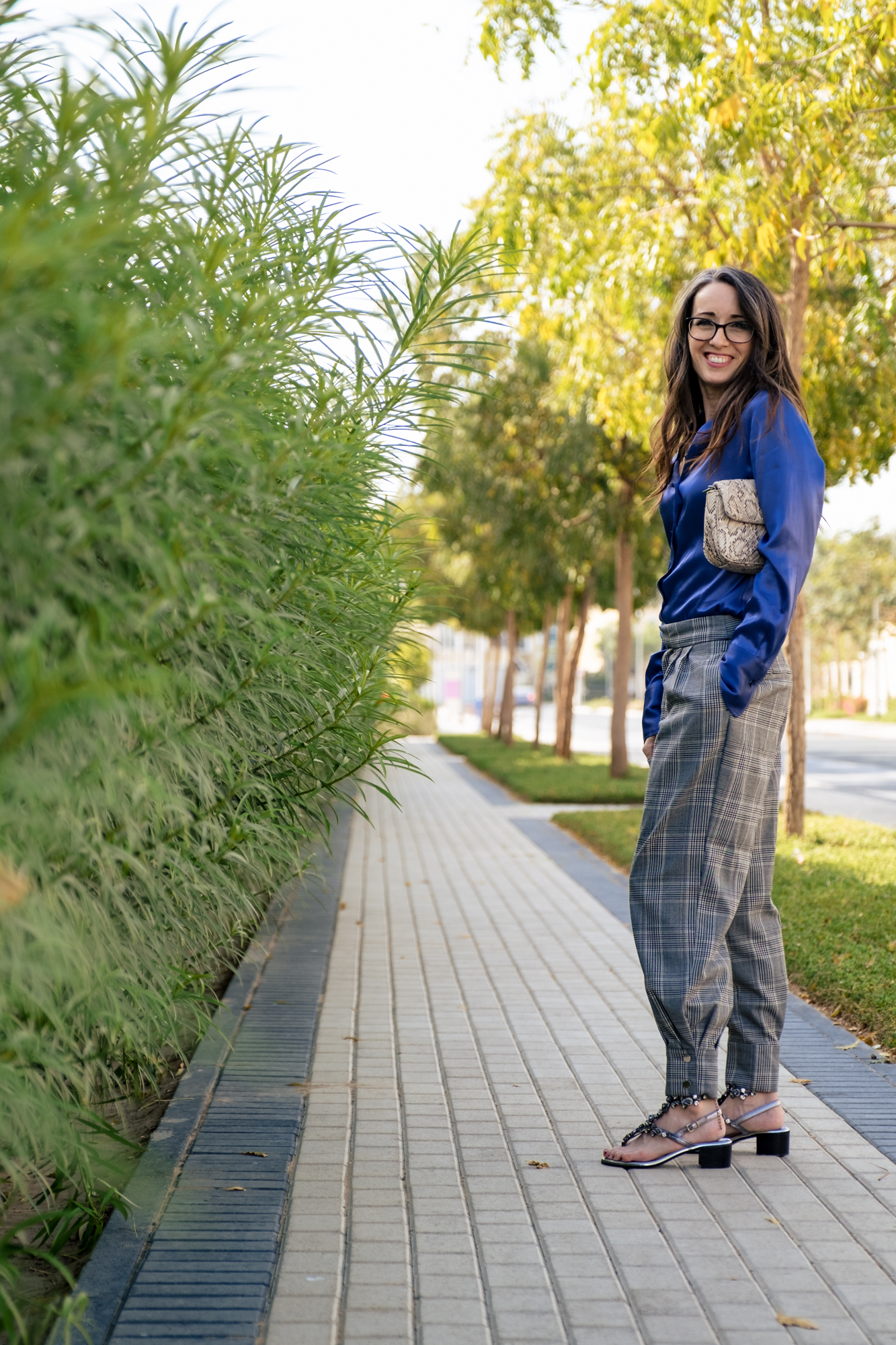 standing to the side, smiling at the camera, blue satin blouse and grey plaid pants