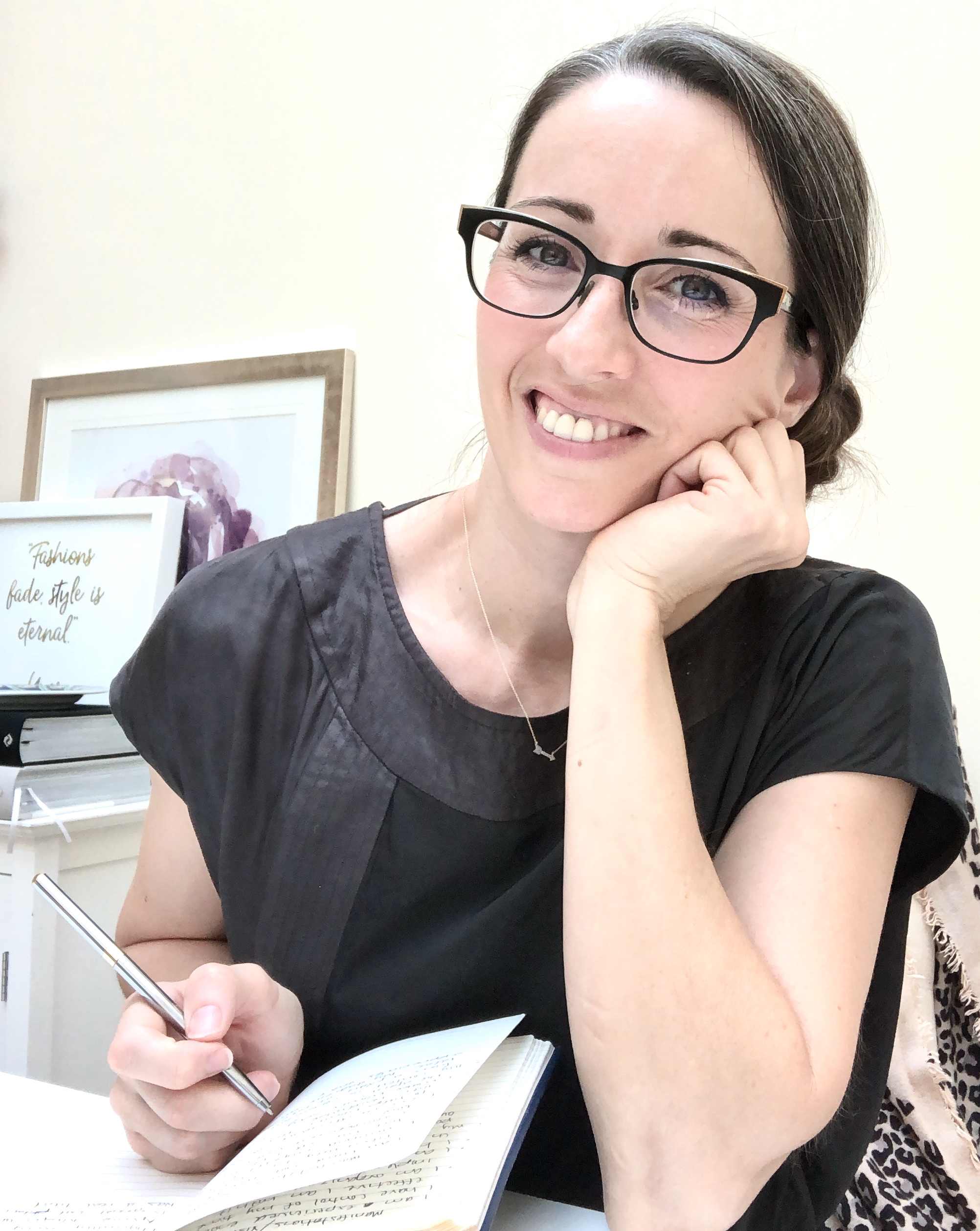 A woman with glasses is smiling and holding her hand on the ear.