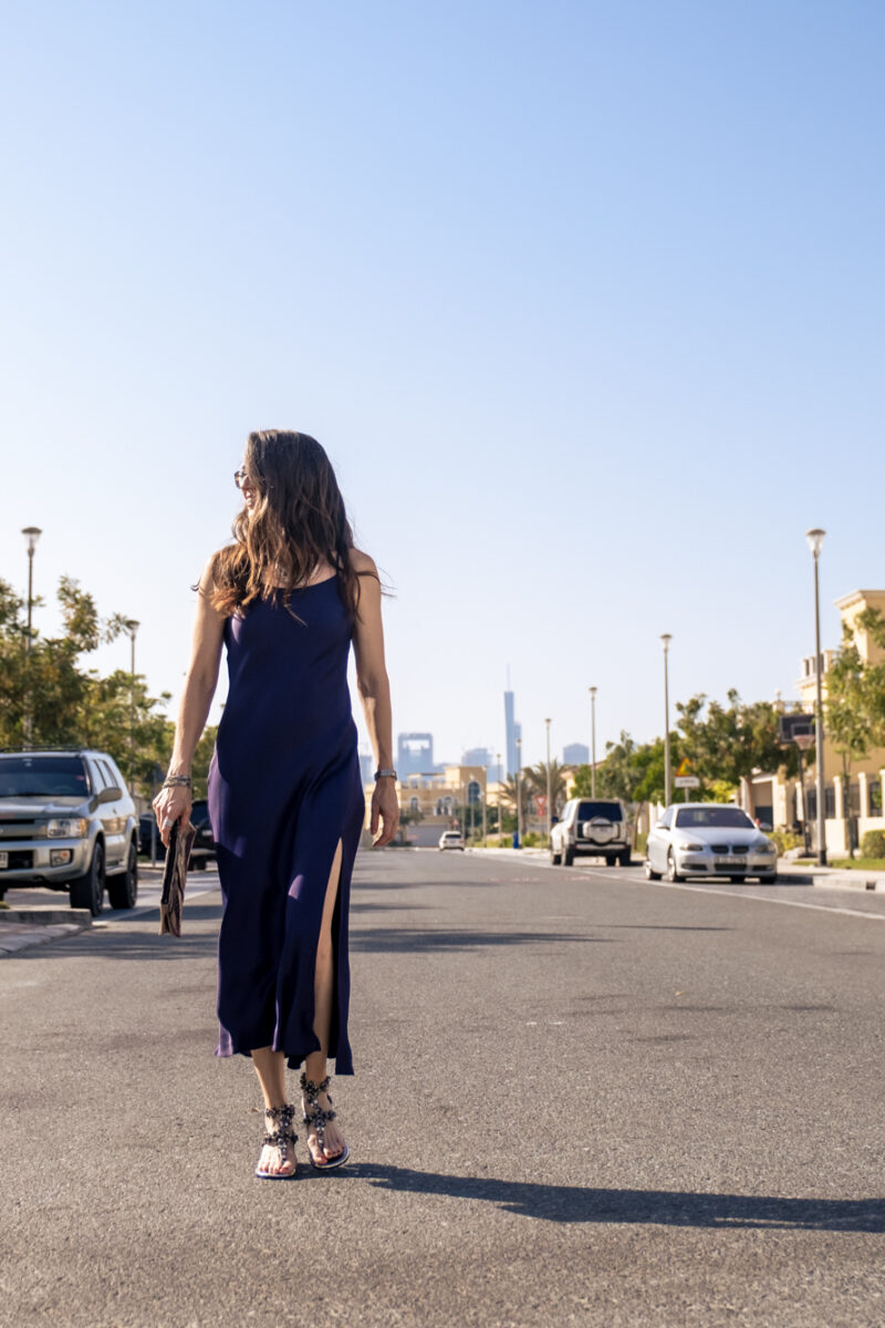 walking on a street towards the camera in a purple dress looking off to the side